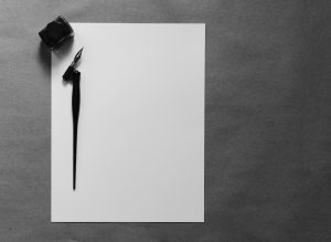 Federhalter mit Tintenfass und leerem Blatt Papier auf hellem Schreibtisch – Symbol für den Schreibbeginn.